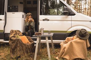 Ein Grand California Van steht geparkt in einem herbstlichen Wald. Eine Frau sitzt in der geöffneten Schiebetür mit einem warmen Getränk, vor dem Van sind ein Campingtisch- und stühle aufgebaut.